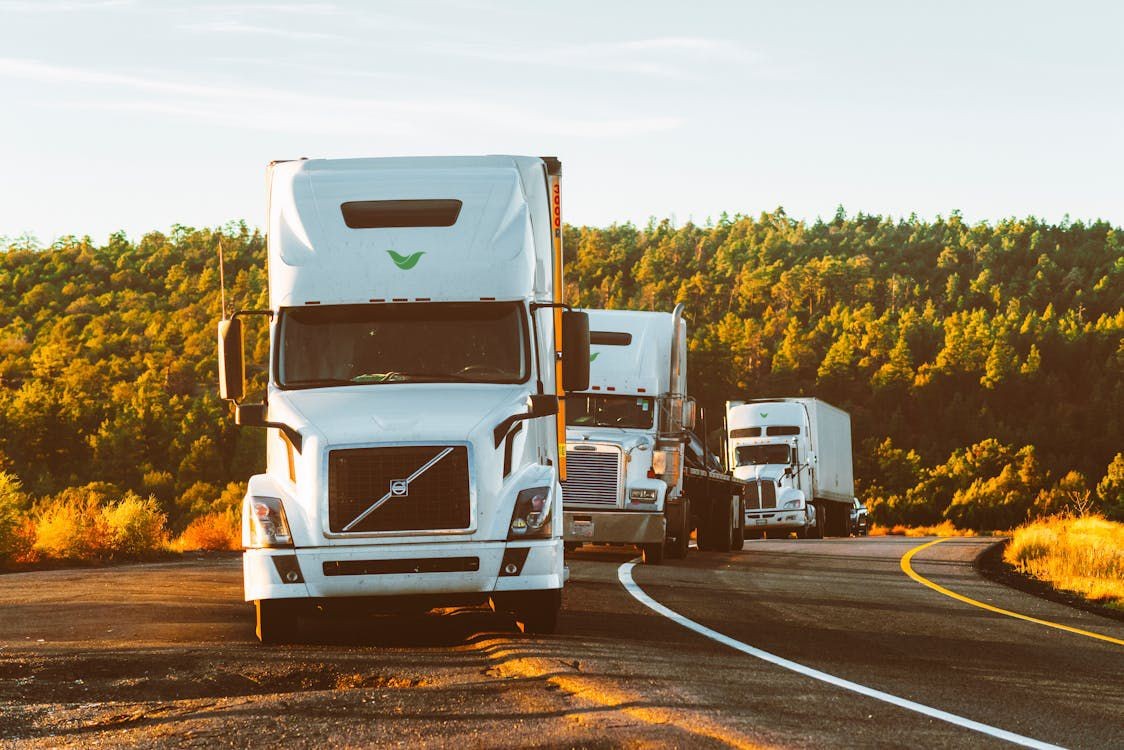 Three white semi-trucks parked on a curved road with a forested background.